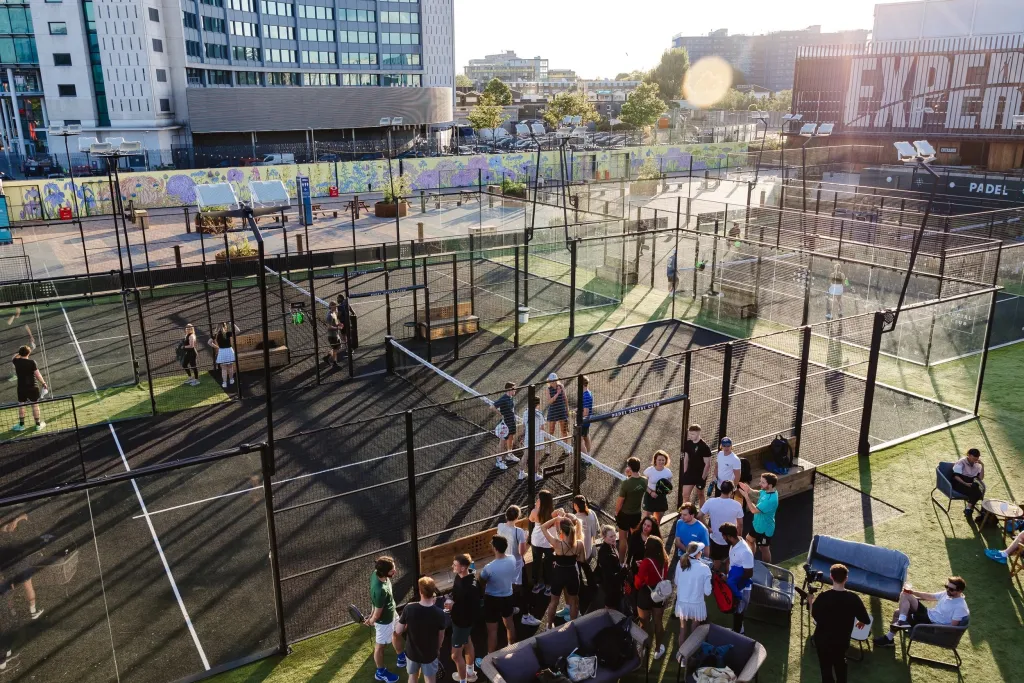 Outdoor padel courts at Padel Social Club in London, with players on court and people socialising on lounge furniture beside the courts